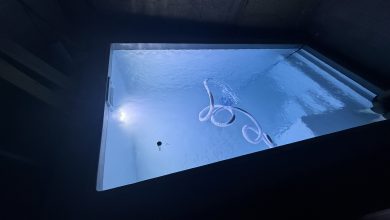 Piscine coque polyester remplie d'eau vue de nuit, éclairée par son projecteur, avec le tuyau flottant du balai en surface, murs du chantier encore bruts autour