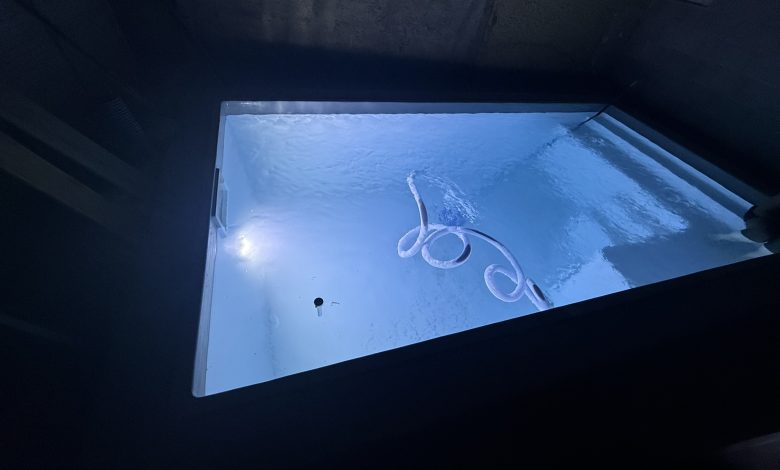 Piscine coque polyester remplie d'eau vue de nuit, éclairée par son projecteur, avec le tuyau flottant du balai en surface, murs du chantier encore bruts autour